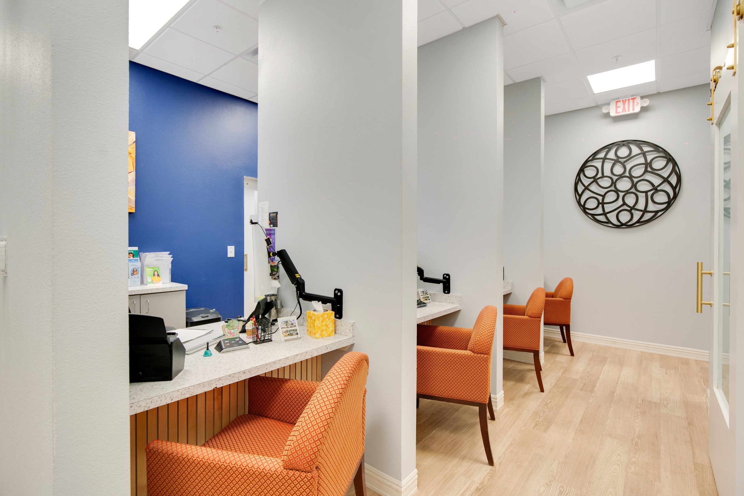 A row of small office or reception cubicles with orange upholstered chairs at desks along light gray walls.